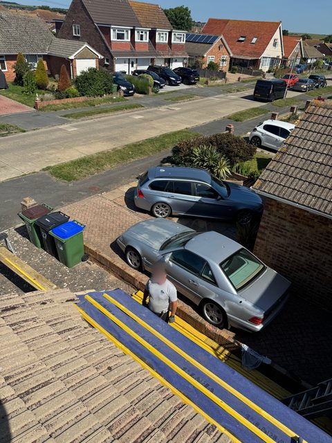 Storm Dry Roofing re-roof: new blue membrane and yellow battens laid down one slope, with a Storm Dry Roofing team member on the driveway below